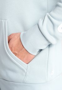 Sweatshirt bleu clair avec une poche kangourou, des poignets côtelés et un petit détail de logo sur le bas devant, présentant un tissu doux et texturé.