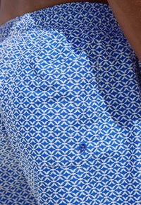 Blå badebukser med mønster, der forestiller bølger i hvid, med elastik i taljen og en enkelt knaplukket lomme.