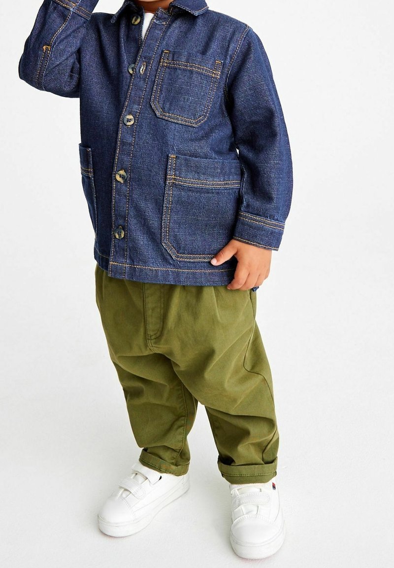 Enfant portant une veste en denim bleu, un pantalon vert olive et des baskets blanches, debout devant un fond blanc uni.