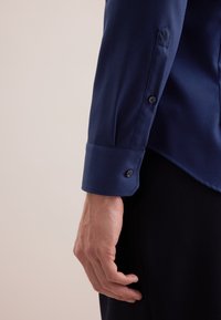 Marineblauw overhemd met knopen van een gestructureerde stof; heeft een omvouwbare manchett en een contrasterende zwarte knoopaccent nabij de mouw.