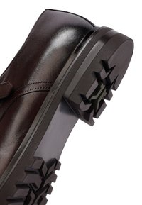 Donkerbruinleren schoenen met een gepolijste afwerking en een dikke zwarte profielzool met diepe groeven voor grip, schuin tegen een witte achtergrond.