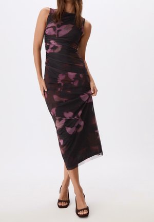 Kobieta w czarnej sukience midi bez rękawów z ciemnofioletowym kwiatowym wzorem, trzymająca dół sukienki, w połączeniu z czarnymi sandałami na otwartym palcu na białym tle.