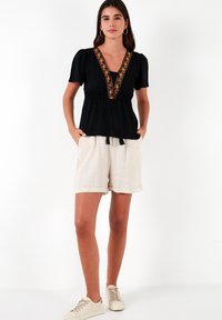 Zwarte top met een V-hals en oranje borduursel, korte pofmouwen en een rekbare taille, gecombineerd met beige shorts en sportschoenen.