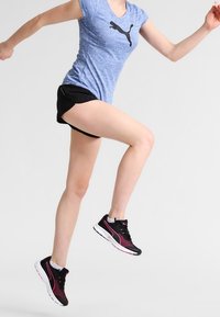 Mujer en medio de un salto, vistiendo zapatillas Puma negras, pantalones cortos negros y una camiseta Puma azul, contra un fondo claro y liso.