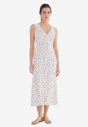Robe blanche à pois noirs, sans manches, avec un col à volants, mi-longue, en tissu léger, portée avec des sandales dorées.