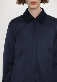Marineblå jakke med spidse krave, tekstilstruktur, forlommer og minimalistisk design, der har en skjult knaplukning.