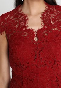 Femme portant une robe en dentelle rouge avec des motifs floraux et un décolleté en V festonné.