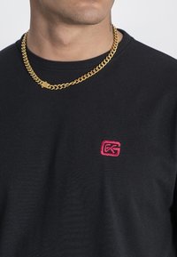 Černé bavlněné tričko s kulatým výstřihem, s malým růžovým vyšitým logem na levé části hrudi. Doplňeno silným zlatým náhrdelníkem.