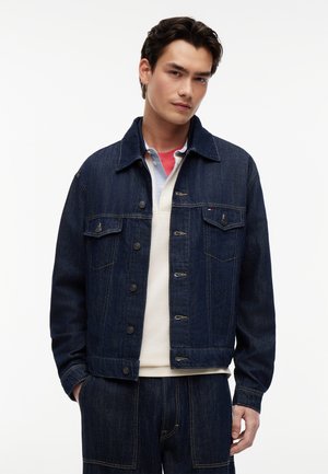 Ung mand iført en mørkeblå denimjakke og matchende bukser, lag på en hvid og rød skjorte, stående mod en hvid baggrund.