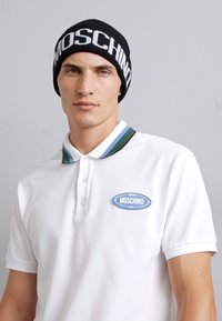 Svart stickad mössa med "MOSCHINO"-logotyp, bärs med en vit polo som har en blå oval logotyp och en grönrandig krage.