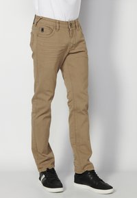 Chinos beige ajustados con una textura suave, que cuentan con bolsillos frontales y un cierre de botón. Combinados con zapatillas negras con cordones.