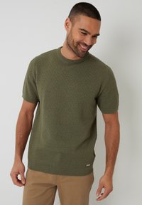 Olijfgroene gebreide trui met korte mouwen en een gestructureerd patroon, ronde hals en ribgebreide manchetten. Draagt bij beige broek.
