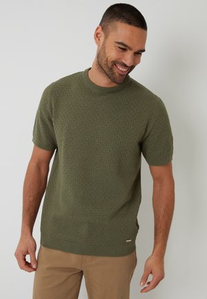 Suéter de punto verde oliva de manga corta con un patrón texturizado, escote redondo y puños de canalé. Llevado con pantalones beige.