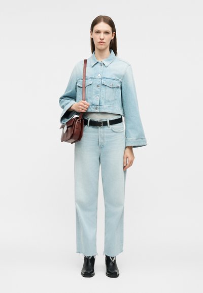 Γυναίκα που στέκεται φορώντας ένα ελαφρύ μπλε cropped τζιν μπουφάν, τζιν ψηλόμεσο, μαύρες μπότες, μαύρη ζώνη και κρατώντας καφέ τσάντα ώμου.