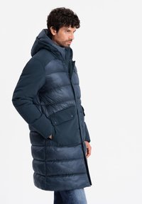 Marineblauer Puffer-Mantel mit Kapuze, der eine Mischung aus matten und glänzenden Texturen, Druckknopf-Taschen und einem Reißverschluss vorne aufweist.