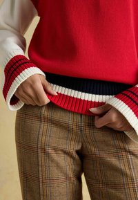 Roter gestrickter Pullover mit breiten weiß-rot gestreiften Bündchen, kombiniert mit braunen karierten Hosen, die gedämpfte blaue und rote Akzente aufweisen.