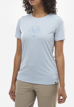 Femme portant une chemise à manches courtes bleu clair avec un motif de montagne et un pantalon beige, debout devant un fond blanc.