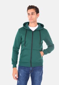Felpa verde con zip in misto cotone, dotata di cappuccio regolabile, tasche frontali e polsini a costine. Indossata sopra una T-shirt bianca, insieme a jeans blu.