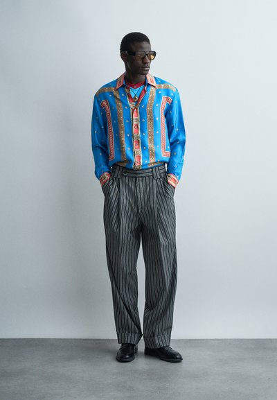 Muž stojaci s rukami v kapse, oblečený v modro-červenej vzorovanej košeli, čierno-šedých nohaviciach s tenkými pruhmi, čiernych topánkach a slnečných okuliaroch.