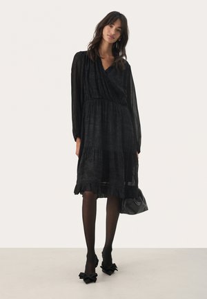 Schwarzes, knielanges Wickelkleid aus transparenter Stoff mit einem volantierten Saum, langen Ärmeln und tailliertem Schnitt. Getragen mit schwarzen Schuhen mit Schleifenakzenten.