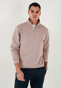 Sudadera ligera de color marrón claro con cremallera y cuello alto, de mangas largas y puños acanalados, usada sobre una camisa blanca, contra un fondo liso.