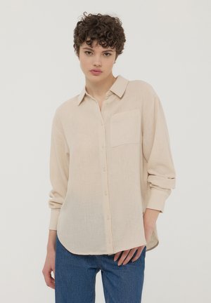 Camicia button-up color beige chiaro con colletto, taschino singolo sul petto e maniche lunghe, realizzata in un tessuto texture. Indossata con jeans blu.