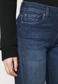 Jeans en denim bleu foncé avec une texture lisse, présentant une taille mi-haute, des poches avant et une décoloration subtile le long de la cuisse.