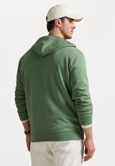 Grön bomullströja med en stor huva, ribbade ärmslut och en texturerad fåll. Bärs med ljusa byxor. Bakifrån visas en enkel design.