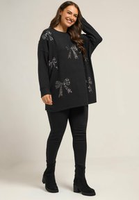 Schwarzer, oversized Pullover mit Pailletten-Schleifen-Design, langen Ärmeln und abgerundetem Ausschnitt. Kombiniert mit schwarzen Slim-Fit-Hosen und Ankle Boots.