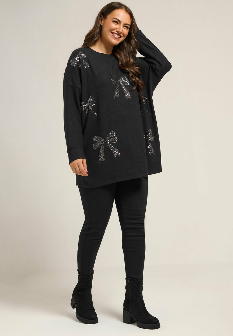Schwarzer, oversized Pullover mit Pailletten-Schleifen-Design, langen Ärmeln und abgerundetem Ausschnitt. Kombiniert mit schwarzen Slim-Fit-Hosen und Ankle Boots.