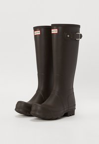 Botas altas de goma marrones con punta redondeada, suela texturizada y detalle de correa ajustable en el lateral. Logotipo de la marca en rojo y blanco en la parte frontal.