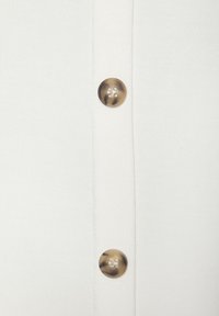 Valge kangast top, millel on vertikaalne rida suuri, ümmarguseid metallnuppe, millel on tekstureeritud pind ja nähtavad õmblused.