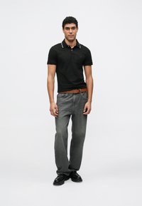 Μαύρο πόλο μπλουζάκι με έντονη γιακά, γκρι τζιν και μαύρα παπούτσια. Το μοντέλο στέκεται με αυτοπεποίθηση μπροστά σε απλό λευκό φόντο.