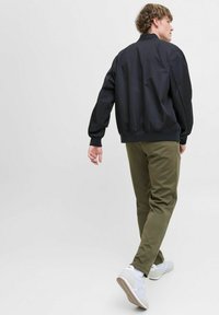 Veste bomber noire avec des poignets côtelés, pantalon tapered vert olive et chaussures de sport blanches. Texture lisse et coupe décontractée, vue de dos.