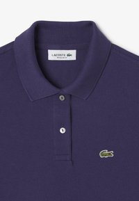 Polo de color púrpura oscuro con una tela texturizada, abertura con dos botones y un pequeño logo de cocodrilo verde en el lado izquierdo del pecho. Diseño de ajuste regular.