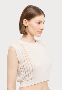 Nauhaton lyhytneulepaita vaaleassa beige-värissä, jossa on teksturoitu kuvio ja avonaiset neuleyksityiskohdat sivuilla. Pyöreä pääntie ja vartalonmyötäinen helma.