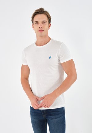 Hvit kortermet t-skjorte laget av mykt stoff, med en liten blå fuglelogo på venstre bryst. Kombinert med mørkeblå jeans.