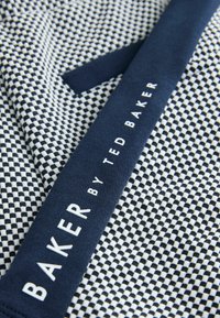 Zbliżenie na czarno-białą tkaninę w kratę z granatowym paskiem z napisem "BAKER BY TED BAKER" białymi literami.