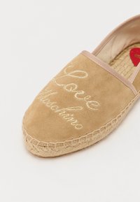 Šviesiai ruda suede espadrilės su auksiniu išsiuvinėtu tekstu "Love Moschino". Yra jūros virvės padas ir kontrastinga audinio apdaila.