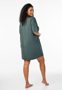 Pārspīlēti liels zaļš kokvilnas kleita ar īsām piedurknēm, gluda tekstūra un vienkāršs dizains, apakšā apmales. Nēsāta basām kājām, skatoties no aizmugures.