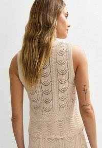 Lace-knit, sleeveless top in light beige, featuring a wave pattern and ribbed texture. Approximately knee-length with scalloped hem details.