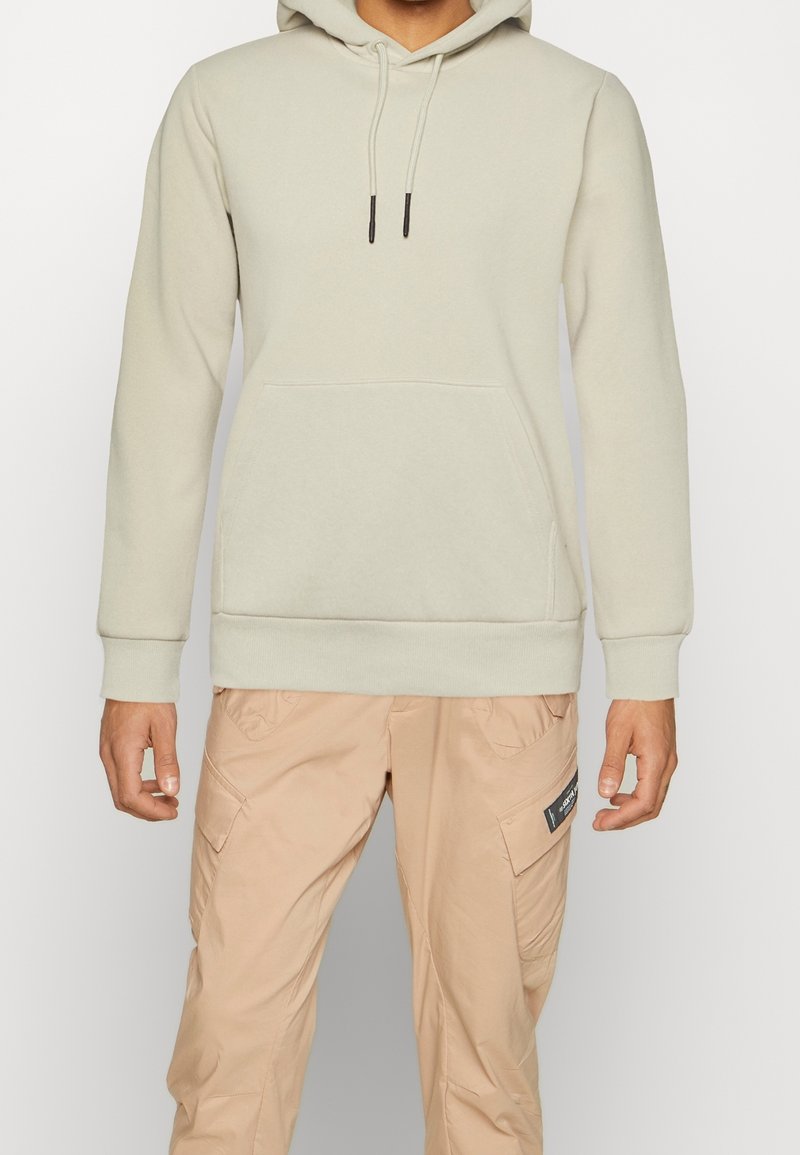 Sudadera gris claro con bolsillo canguro, puños acanalados y capucha con cordón. Combinada con pantalones cargo beige claros que tienen bolsillos laterales.