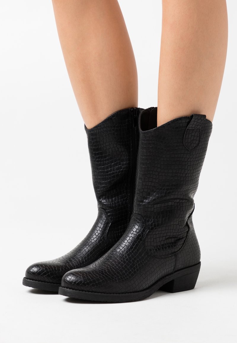 Bottes noires mi-mollet à texture crocodile portées sur des jambes nues, avec des talons bas et des fermetures éclair latérales, sur un fond blanc.