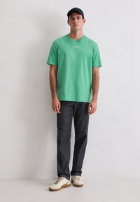 Zielony bawełniany t-shirt z krótkim rękawem, szare spodnie i kremowe trampki. T-shirt ma na przodzie małe logo tekstowe. Brak wzorów i akcentów.