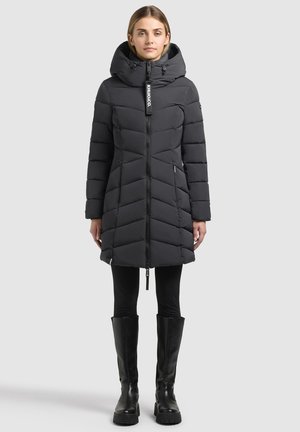 Femme portant un long manteau d'hiver noir matelassé avec un col montant, des leggings noirs et des bottes noires montantes sur un fond clair uni.
