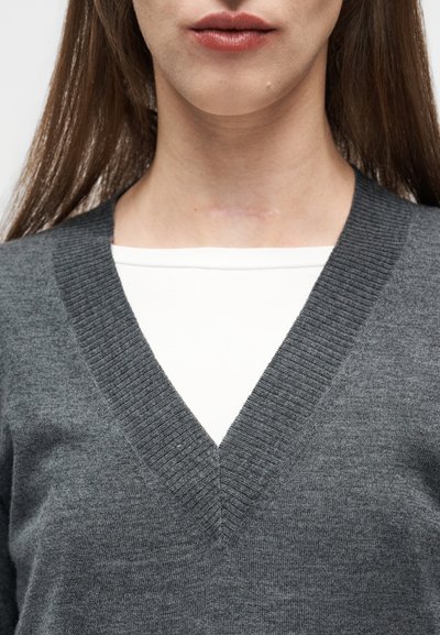 Νεαρή γυναίκα που φοράει γκρι πουλόβερ με V λαιμόκοψη και έχει εμφανές σημάδι στο μπροστινό μέρος του λαιμού κάτω από το πηγούνι.