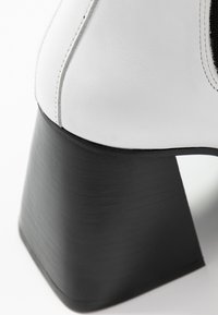 Bota de tobillo blanca de cuero con un tacón geométrico negro, que presenta una textura suave y un detalle de costura a lo largo de la costura.