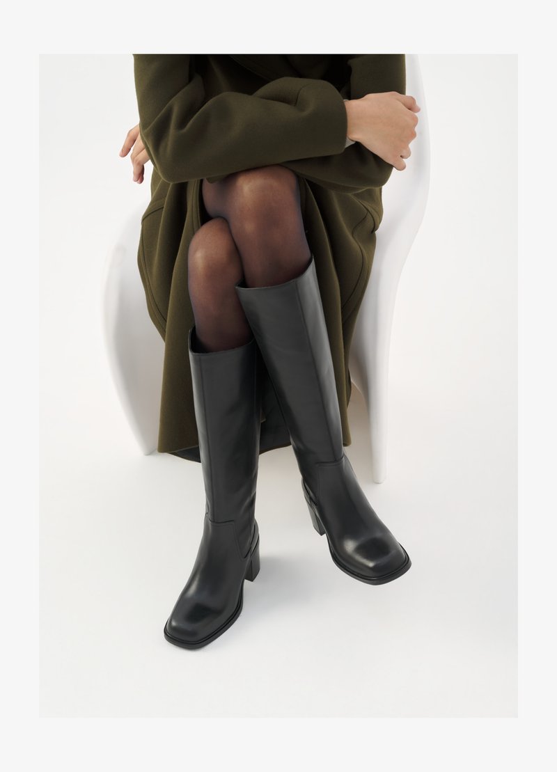 Térdfelett érő fekete bőr csizmák négyzet alakú orral és tömbtalppal, olajzöld kabátban ülő modellel bemutatva. Simaság, minimalista részletezés.