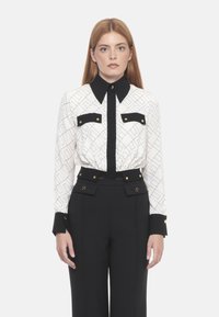 Blusa bianca e nera a fantasia con colletto a punta e bottoni dorati, abbinata a pantaloni neri a vita alta con dettagli a bottoni.