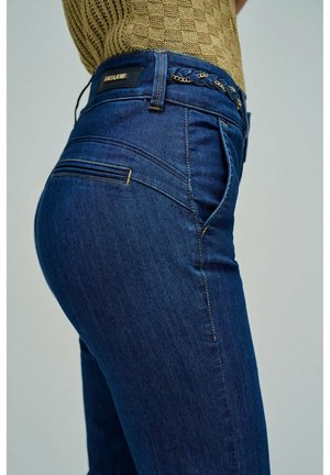 Persona con vaqueros ajustados azul oscuro, cinturón trenzado y camiseta de manga corta texturizada color marrón claro, vista lateral de la cintura al muslo.